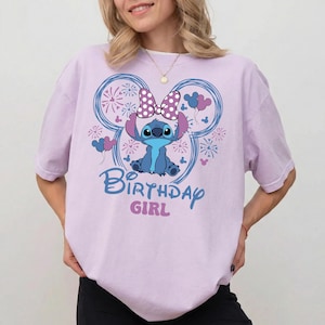 May include: A light purple t-shirt featuring a cartoon character with a polka dot bow, surrounded by fireworks and balloons. The text "Birthday Girl" is printed below the character in blue and pink.