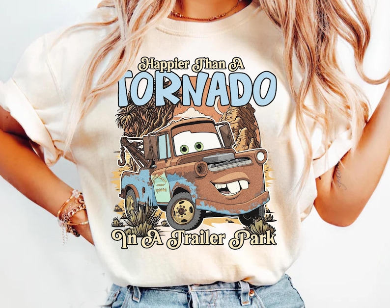 Op de afbeelding: Een cr&egrave;mekleurig t-shirt met een afbeelding van een roestige bruine en blauwe takelwagen met groene ogen en een glimlach met een spleetje tussen de tanden. De tekst Happier Than A TORNADO In A Trailer Park staat voor een woestijnlandschap.