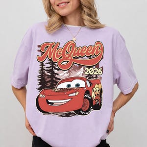 Puede incluir: Una camiseta de manga corta de color morado claro con un estampado gr&aacute;fico de un coche de carreras rojo con el n&uacute;mero 95. Sobre el coche, el texto dice McQueen en una fuente estilizada, con el a&ntilde;o 2026 impreso a la derecha.