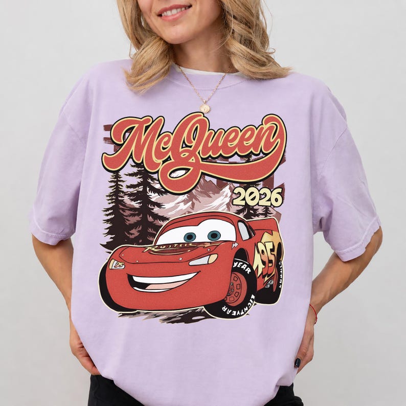 Puede incluir: Una camiseta de manga corta de color morado claro con un estampado gr&aacute;fico de un coche de carreras rojo con el n&uacute;mero 95. Sobre el coche, el texto dice McQueen en una fuente estilizada, con el a&ntilde;o 2026 impreso a la derecha.