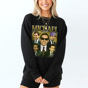 May include: Black sweatshirt featuring a collage of images of Michael Scott, with the name "MICHAEL Scott" in gold lettering. The images show Scott in various expressions and outfits. The sweatshirt is a crew neck style.