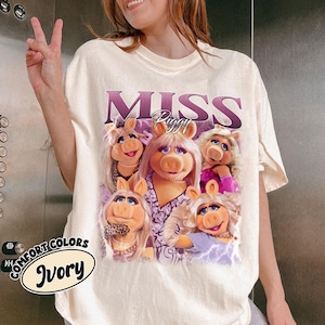 May include: Ivory-colored t-shirt featuring a graphic of Miss Piggy and the text "MISS PIGGY" in purple. The graphic includes multiple images of Miss Piggy. The shirt also has the words "COMFORT COLORS" and "Ivory" printed on it.
