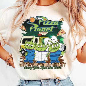 May include: A cream-colored t-shirt featuring a graphic print of three green three-eyed aliens. The design includes the text Pizza Planet, Out of This World Quality, and Serving Your Local Star Cluster, along with a yellow delivery truck and pizza slices.