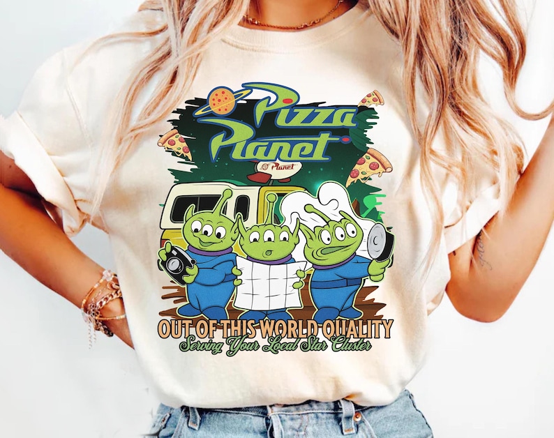 May include: A cream-colored t-shirt featuring a graphic print of three green three-eyed aliens. The design includes the text Pizza Planet, Out of This World Quality, and Serving Your Local Star Cluster, along with a yellow delivery truck and pizza slices.
