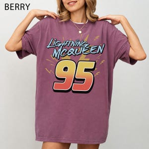 May include: A berry-coloured short-sleeve t-shirt featuring a graphic print with the text Lightning McQueen above the number 95 in a yellow-to-red gradient. The design includes yellow lightning bolt accents on a solid purple-toned fabric.