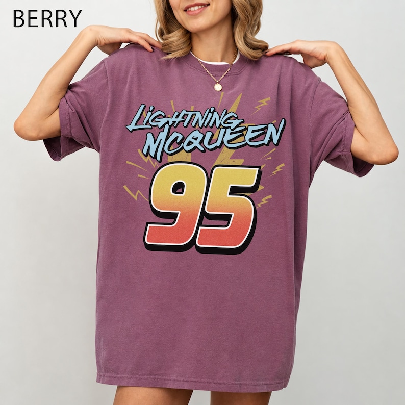 May include: A berry-coloured short-sleeve t-shirt featuring a graphic print with the text Lightning McQueen above the number 95 in a yellow-to-red gradient. The design includes yellow lightning bolt accents on a solid purple-toned fabric.