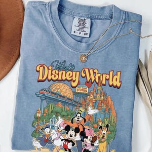 Pode incluir: Uma t-shirt azul de gola redonda com uma estampa gr&aacute;fica do Walt Disney World, apresentando um monotrilho, um castelo e personagens como Mickey Mouse, Minnie Mouse, Donald Duck, Daisy Duck, Goofy e Pluto. Possui uma etiqueta Comfort Colors.
