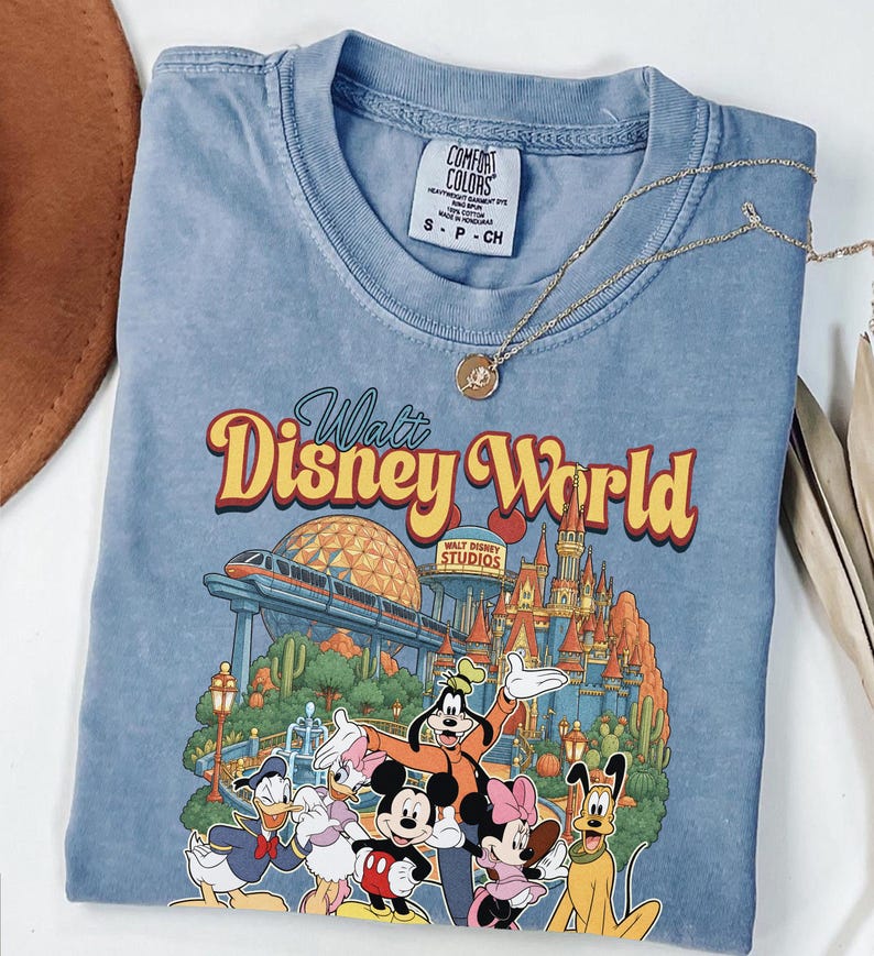 Pode incluir: Uma t-shirt azul de gola redonda com uma estampa gr&aacute;fica do Walt Disney World, apresentando um monotrilho, um castelo e personagens como Mickey Mouse, Minnie Mouse, Donald Duck, Daisy Duck, Goofy e Pluto. Possui uma etiqueta Comfort Colors.