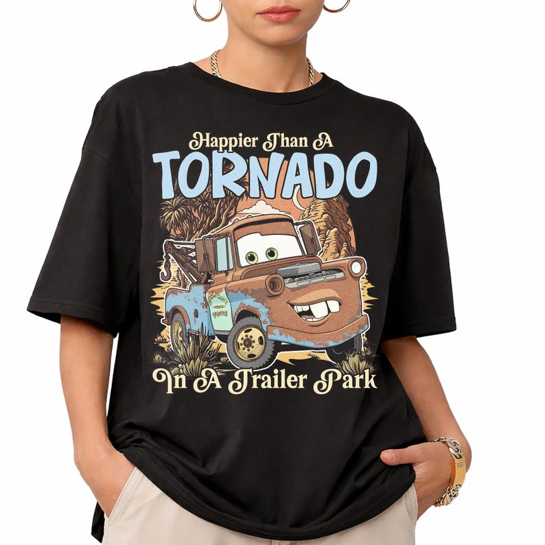 Op de afbeelding: Een zwart t-shirt met korte mouwen en een afbeelding van een roestbruine takelwagen met grote ogen en een brede glimlach. De tekst erboven luidt Happier Than A TORNADO en de tekst eronder In A Trailer Park, tegen een woestijnlandschap.