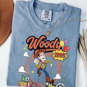 May include: A blue short-sleeved t-shirt featuring a graphic of the character Woody from Toy Story. The design includes the text Woody and 1995, colorful balloons, alphabet blocks, clouds, stars, and a sheriff badge. The shirt has a Comfort Colors label.