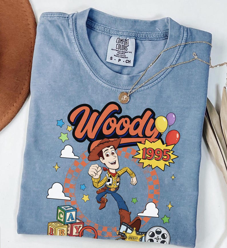 May include: A blue short-sleeved t-shirt featuring a graphic of the character Woody from Toy Story. The design includes the text Woody and 1995, colorful balloons, alphabet blocks, clouds, stars, and a sheriff badge. The shirt has a Comfort Colors label.