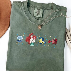 Może przedstawiać: Oliwkowozielony t-shirt z kr&oacute;tkim rękawem z nadrukiem graficznym przedstawiającym Arielkę, Florka, Sebastiana i perłę w muszli, otoczonych małymi ż&oacute;łtymi gwiazdkami i niebieskimi bąbelkami. Koszulka posiada metkę Comfort Colors przy wewnętrznym kołnierzu.