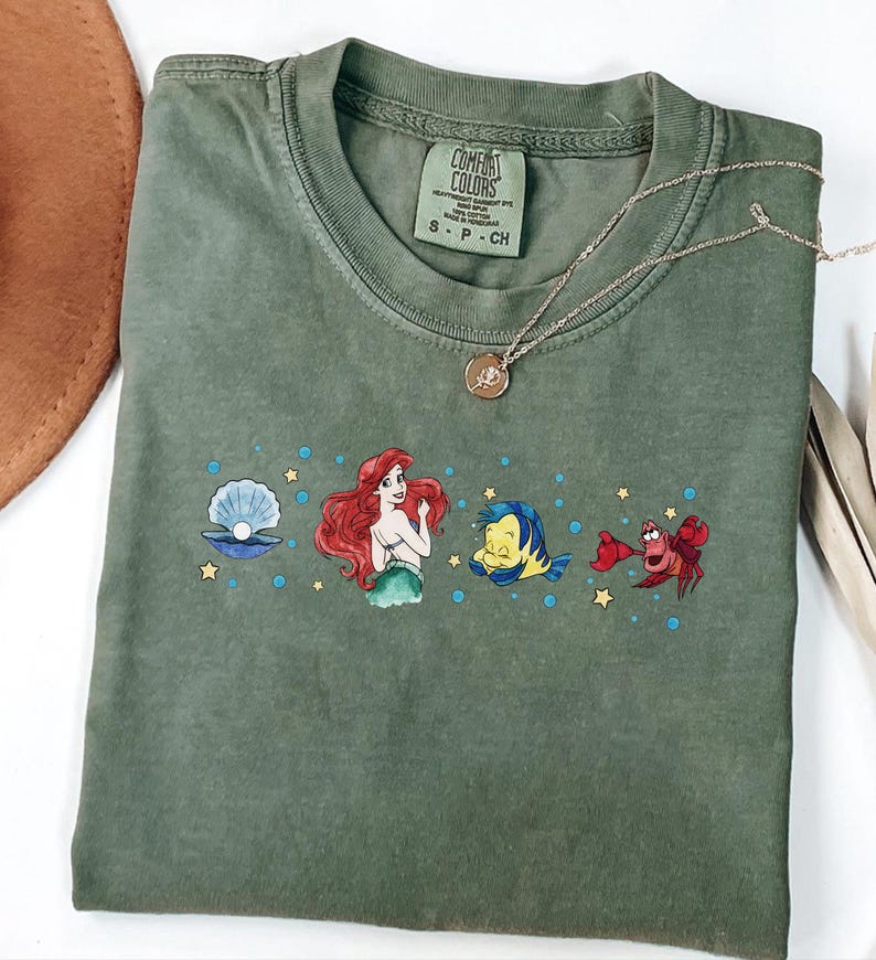 Może przedstawiać: Oliwkowozielony t-shirt z kr&oacute;tkim rękawem z nadrukiem graficznym przedstawiającym Arielkę, Florka, Sebastiana i perłę w muszli, otoczonych małymi ż&oacute;łtymi gwiazdkami i niebieskimi bąbelkami. Koszulka posiada metkę Comfort Colors przy wewnętrznym kołnierzu.