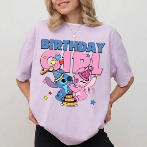 May include: A light purple t-shirt featuring the words "BIRTHDAY GIRL" in blue and pink. The graphic includes cartoon characters Stitch and Angel, with a birthday cake and party hats. Stars and hearts decorate the design.