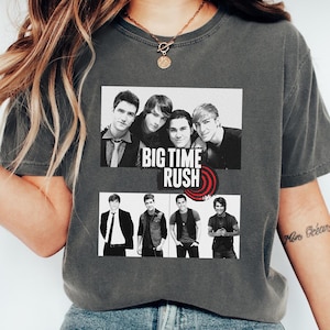 以下が含まれることがあります： ビッグタイムラッシュの白黒グラフィックが特徴の濃いグレーのTシャツ。グラフィックには、バンドメンバーの2つの画像とバンドのロゴが含まれています。シャツは柔らかい素材で作られています。