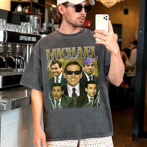 May include: Dark gray t-shirt featuring a collage of images and the name "Michael Scott" in gold lettering. The shirt showcases various images of a man in a suit, with one image wearing sunglasses and another with a purple bandana. The shirt is a vintage style.