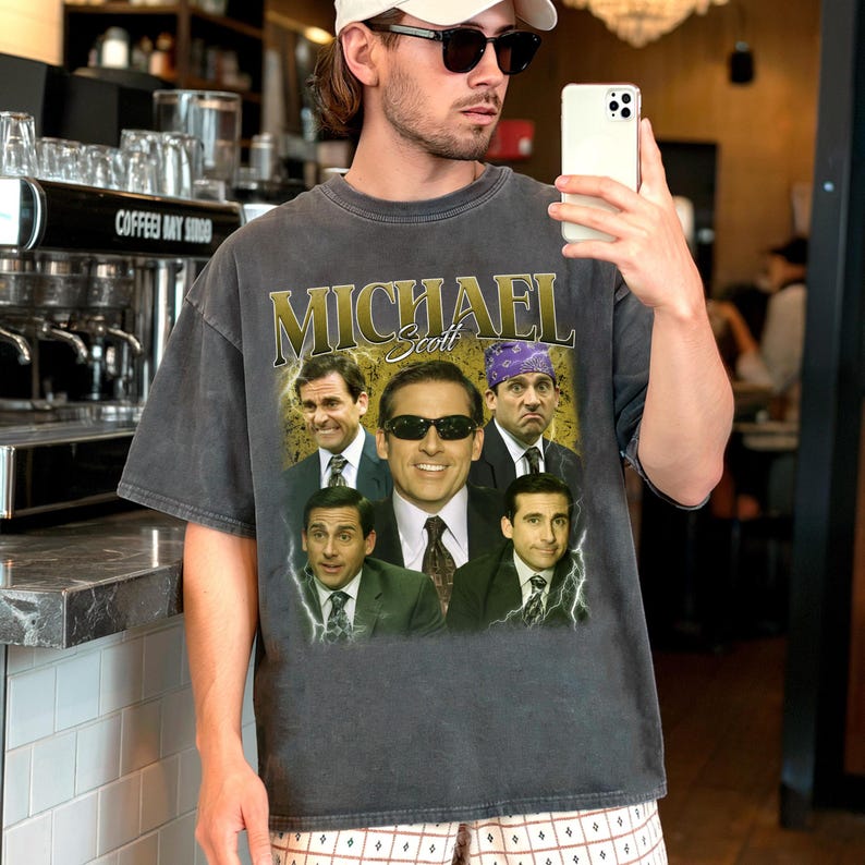 May include: Dark gray t-shirt featuring a collage of images and the name "Michael Scott" in gold lettering. The shirt showcases various images of a man in a suit, with one image wearing sunglasses and another with a purple bandana. The shirt is a vintage style.