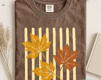 May include: A brown crew neck t-shirt featuring a graphic of three maple leaves in shades of yellow and orange layered over vertical cream-colored stripes. The inner neck label displays the text Comfort Colors.