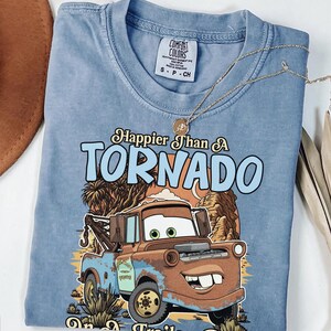 Op de afbeelding: Een blauw t-shirt met korte mouwen en een afbeelding van een roestige bruine takelwagen. De tekst luidt Happier Than A TORNADO on a Trailer Park. Het shirt heeft een Comfort Colors merklabel in de nek en een kleine goudkleurige ketting.