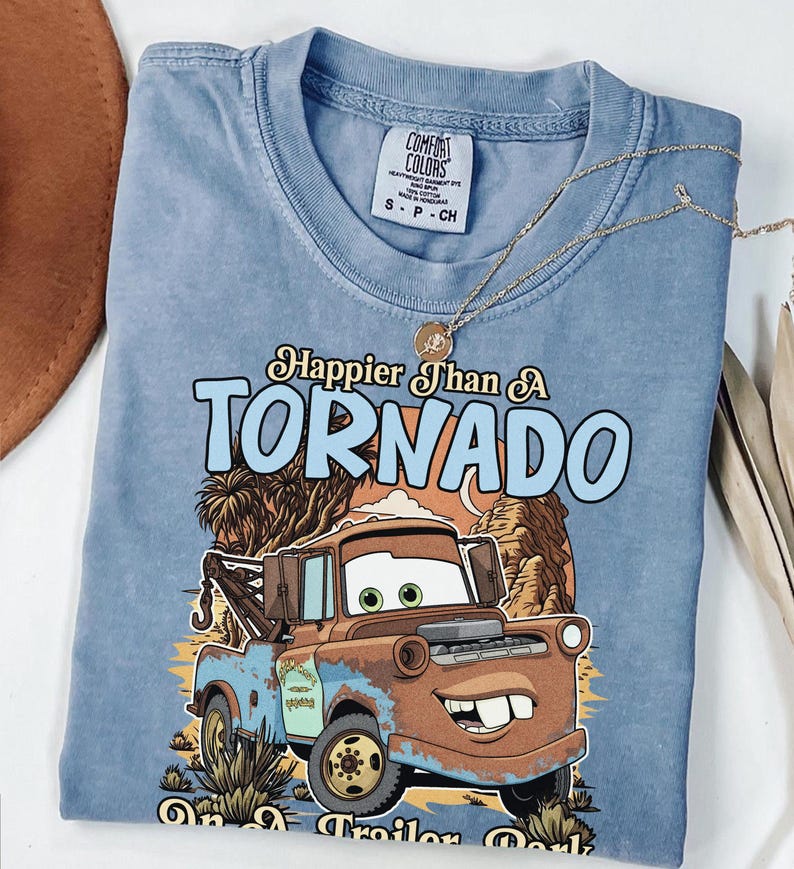 Op de afbeelding: Een blauw t-shirt met korte mouwen en een afbeelding van een roestige bruine takelwagen. De tekst luidt Happier Than A TORNADO on a Trailer Park. Het shirt heeft een Comfort Colors merklabel in de nek en een kleine goudkleurige ketting.