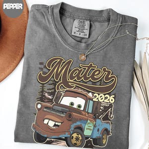 May include: A charcoal gray t-shirt featuring a graphic of a brown and blue tow truck character with large eyes and a wide smile. The text "Mater" is printed in a large brown script font above the truck, with the year "2026" in yellow numerals.