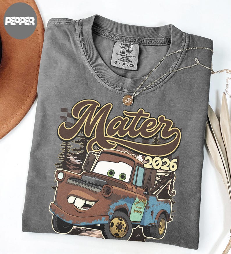 May include: A charcoal gray t-shirt featuring a graphic of a brown and blue tow truck character with large eyes and a wide smile. The text "Mater" is printed in a large brown script font above the truck, with the year "2026" in yellow numerals.