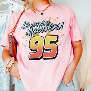 May include: A light pink short-sleeve t-shirt featuring the text Lightning McQueen in blue letters above the large number 95, which has a yellow to orange gradient fill and a black outline, accented by yellow lightning bolt graphic elements.