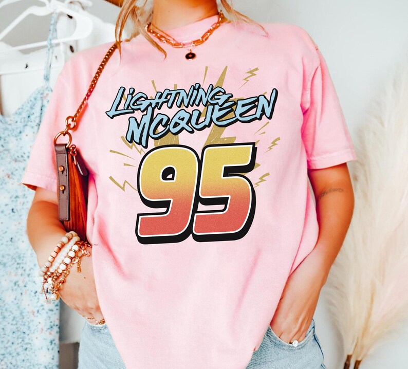 May include: A light pink short-sleeve t-shirt featuring the text Lightning McQueen in blue letters above the large number 95, which has a yellow to orange gradient fill and a black outline, accented by yellow lightning bolt graphic elements.