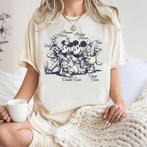 Może przedstawiać: Kremowy t-shirt z krótkim rękawem z ciemnogranatową grafiką przedstawiającą Goofy'ego, Minnie Mouse, Mickey Mouse, Daisy Duck, Pluto i Donalda Ducka, z ich imionami wydrukowanymi kursywą nad i pod każdą z postaci.