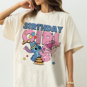 May include: Cream-colored t-shirt featuring the text "BIRTHDAY GIRL" in blue and pink. The graphic includes cartoon characters Stitch and Angel, a birthday cake, and star and heart accents. The shirt has short sleeves.