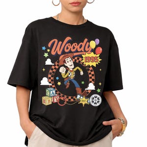 May include: A black t-shirt featuring a graphic of the character Woody from Toy Story. The design includes the text Woody and 1995, a checkered circle, colorful stars, clouds, alphabet blocks, a sheriff badge, and a film reel on the front.