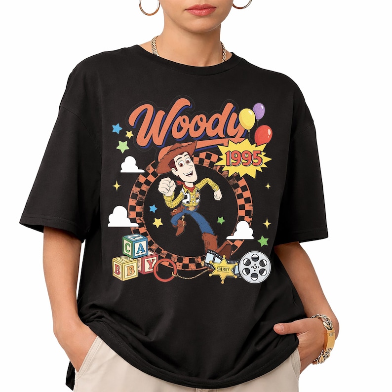 May include: A black t-shirt featuring a graphic of the character Woody from Toy Story. The design includes the text Woody and 1995, a checkered circle, colorful stars, clouds, alphabet blocks, a sheriff badge, and a film reel on the front.