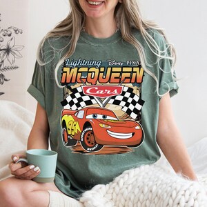 May include: A sage green short-sleeve t-shirt featuring a graphic of Lightning McQueen from Disney Pixar Cars. The design includes the text Lightning McQueen, a red race car with the number 95, and black and white checkered racing flags.