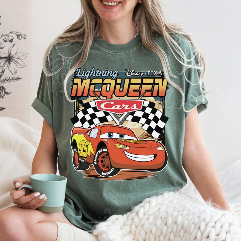 May include: A sage green short-sleeve t-shirt featuring a graphic of Lightning McQueen from Disney Pixar Cars. The design includes the text Lightning McQueen, a red race car with the number 95, and black and white checkered racing flags.