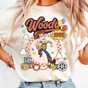 May include: A cream-colored t-shirt featuring a graphic print of the character Woody from Toy Story. The design includes the text Woody and 1995, colorful balloons, alphabet blocks, a sheriff badge, and a film reel against a circular background.
