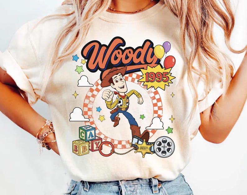 May include: A cream-colored t-shirt featuring a graphic print of the character Woody from Toy Story. The design includes the text Woody and 1995, colorful balloons, alphabet blocks, a sheriff badge, and a film reel against a circular background.