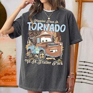 Op de afbeelding: Een antracietgrijs T-shirt met korte mouwen en een afbeelding van een roestbruine takelwagen met grote ogen en een brede glimlach. De tekst luidt Happier Than A TORNADO In A Trailer Park in lichtblauwe en cr&egrave;mekleurige letters.