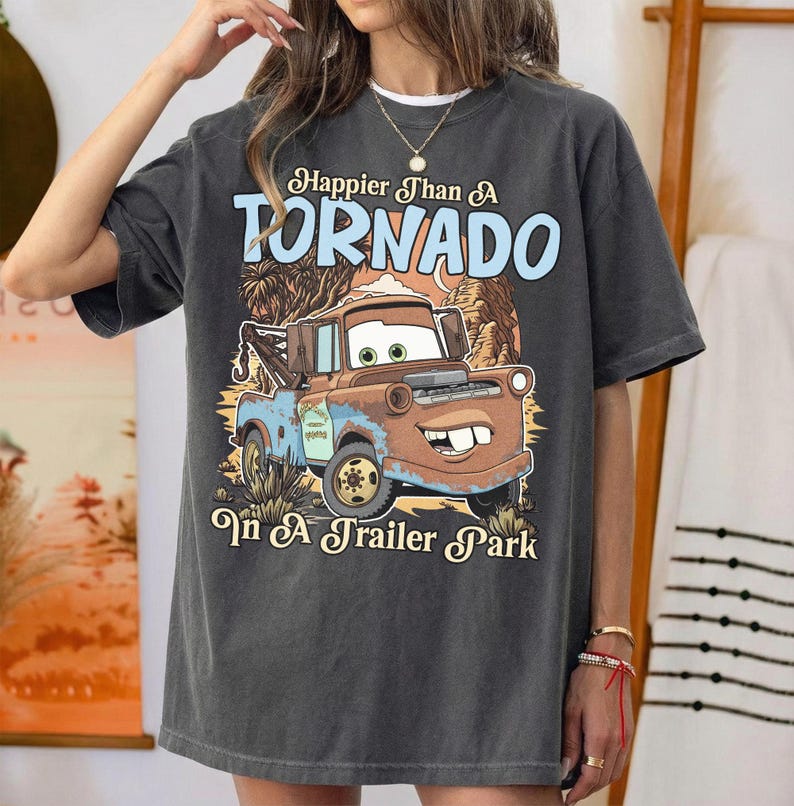 Op de afbeelding: Een antracietgrijs T-shirt met korte mouwen en een afbeelding van een roestbruine takelwagen met grote ogen en een brede glimlach. De tekst luidt Happier Than A TORNADO In A Trailer Park in lichtblauwe en cr&egrave;mekleurige letters.
