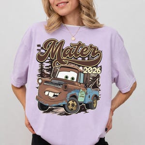 May include: A light purple t-shirt featuring a graphic print of a brown and blue tow truck character with large eyes and buck teeth. The text "Mater" appears in a large brown script font above the truck, with the number "2026" printed to the right.