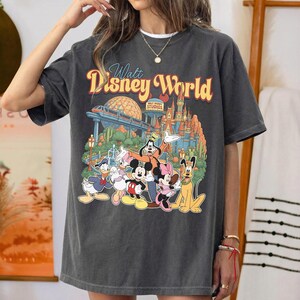 Pode incluir: Uma t-shirt cinza-carv&atilde;o de manga curta com uma estampa gr&aacute;fica do Walt Disney World, apresentando Mickey Mouse, Minnie Mouse, Pato Donald, Margarida, Pateta e Pluto. O design inclui o castelo do parque e a esfera do Epcot ao fundo.