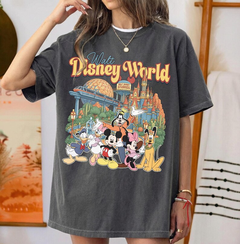 Pode incluir: Uma t-shirt cinza-carv&atilde;o de manga curta com uma estampa gr&aacute;fica do Walt Disney World, apresentando Mickey Mouse, Minnie Mouse, Pato Donald, Margarida, Pateta e Pluto. O design inclui o castelo do parque e a esfera do Epcot ao fundo.