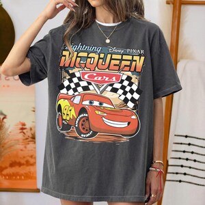 May include: A charcoal gray short-sleeve t-shirt featuring a graphic of Lightning McQueen from the Disney Pixar movie Cars. The design includes the text Lightning McQueen, the Cars logo, two checkered racing flags, and the number 95 on the car.