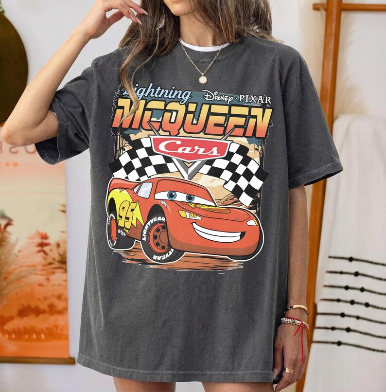 May include: A charcoal gray short-sleeve t-shirt featuring a graphic of Lightning McQueen from the Disney Pixar movie Cars. The design includes the text Lightning McQueen, the Cars logo, two checkered racing flags, and the number 95 on the car.