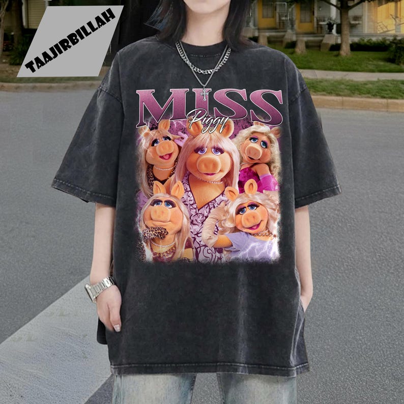 May include: Dark gray t-shirt featuring a graphic of Miss Piggy and the text "MISS Piggy" in pink and purple. The image shows Miss Piggy in various poses. The shirt has short sleeves and a crew neck.