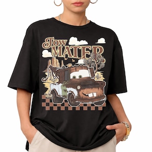 May include: A black t-shirt featuring a graphic print of the character Tow Mater. The design includes the text "Tow Mater" in a stylized font above the rusty brown tow truck, set against a desert landscape with cacti and a checkered pattern.