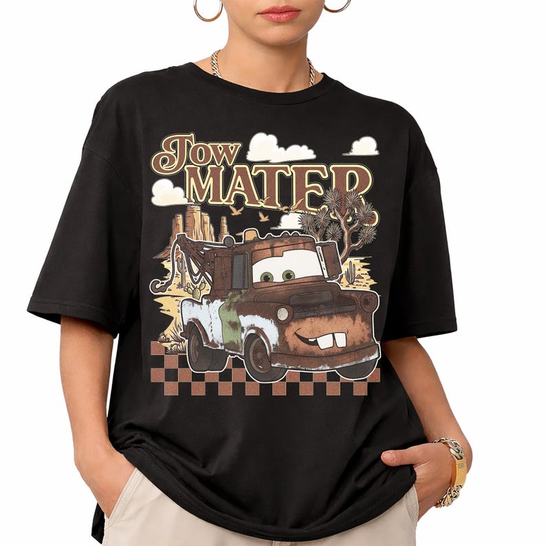 May include: A black t-shirt featuring a graphic print of the character Tow Mater. The design includes the text "Tow Mater" in a stylized font above the rusty brown tow truck, set against a desert landscape with cacti and a checkered pattern.