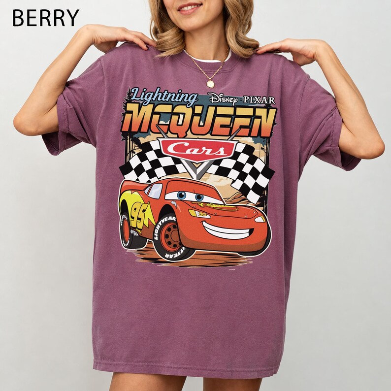 May include: A berry-colored t-shirt featuring a graphic print of Lightning McQueen from the Disney Pixar movie Cars. The design includes the text Lightning McQueen, the Cars logo, and a checkered flag background above the red race car character.