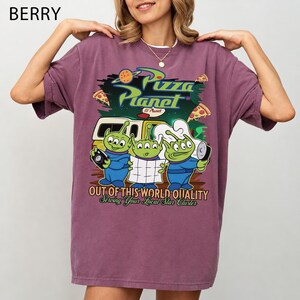 May include: A berry-colored short-sleeve t-shirt featuring a graphic print of three green aliens from Pizza Planet. The design includes the text Pizza Planet, Out of This World Quality, and Serving Your Local Star Cluster with pizza slices.