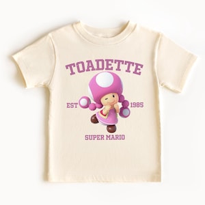 May include: A cream-coloured t-shirt with a graphic of Toadette from Super Mario. The design includes the text "TOADETTE", "EST 1985", and "SUPER MARIO" in purple. The character is shown in a pink dress and mushroom cap.