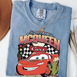 May include: A blue short-sleeve t-shirt featuring a graphic of the red race car Lightning McQueen from the movie Cars. The design includes the text Lightning McQueen, a traffic light, checkered flags, and a cactus. The shirt has a Comfort Colors label.